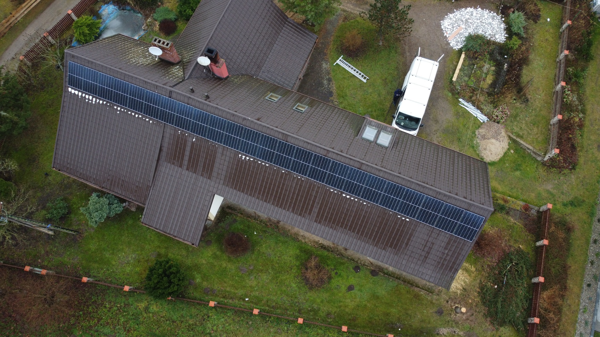 Widok z lotu ptaka na brązowy dach z panelami fotowoltaicznymi. Obok zaparkowany biały bus, a na trawie leży drabina.