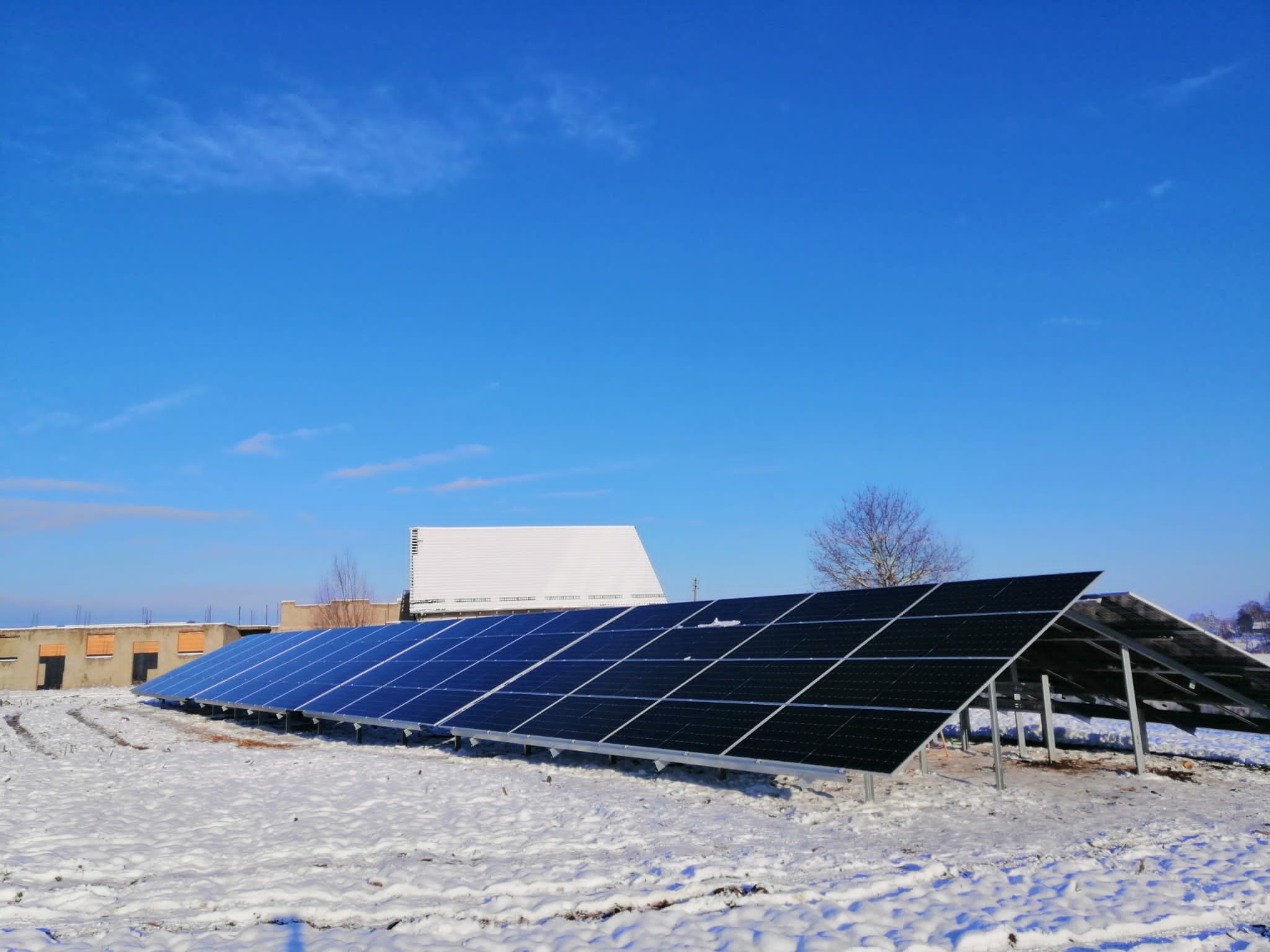 Rząd paneli fotowoltaicznych pokrytych śniegiem na tle błękitnego nieba i częściowo ośnieżonego budynku. Widoczne metalowe konstrukcje wsporcze.