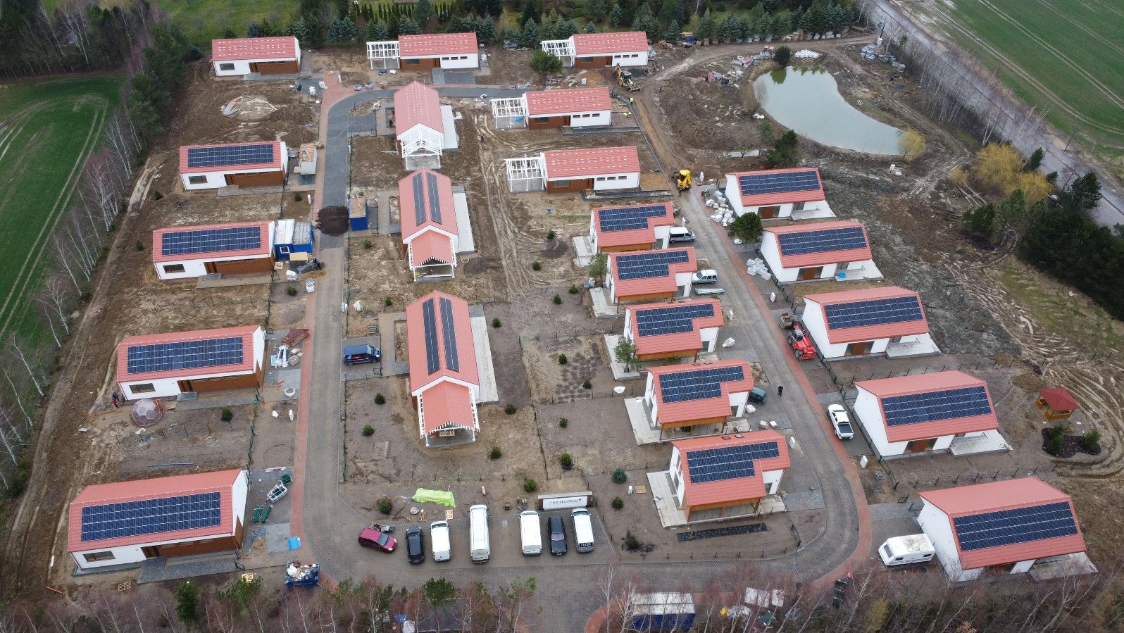 Panoramiczny widok z lotu ptaka na osiedle domów jednorodzinnych z panelami fotowoltaicznymi na dachach, otoczone zielenią i w trakcie zagospodarowania terenu.