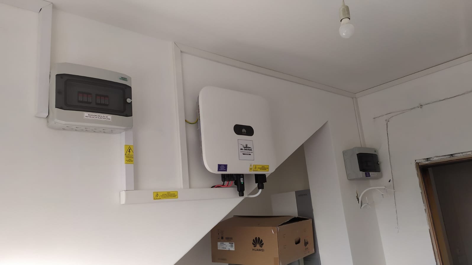 Biała ściana z zamontowaną rozdzielnicą PV DC oraz falownikiem Huawei. Widoczna skrzynka kartonowa z logo Huawei. Całość instalacji wewnątrz budynku.