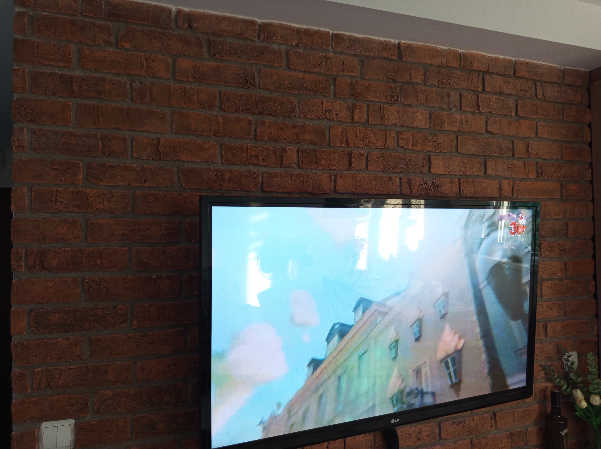 Ściana z cegły dekoracyjnej za telewizorem, widoczny fragment sufitu i listwa przypodłogowa. Styl rustykalny z nowoczesnym akcentem w postaci płaskiego ekranu.
