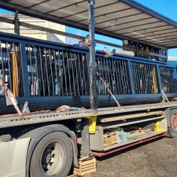 SPEED ROAD Adrian Izydorczyk - Ciężarówka z naczepą przewozi czarne, metalowe elementy ogrodzenia zabezpieczone pasami transportowymi w słoneczny dzień. Widoczne skrzynki i narzędzia.