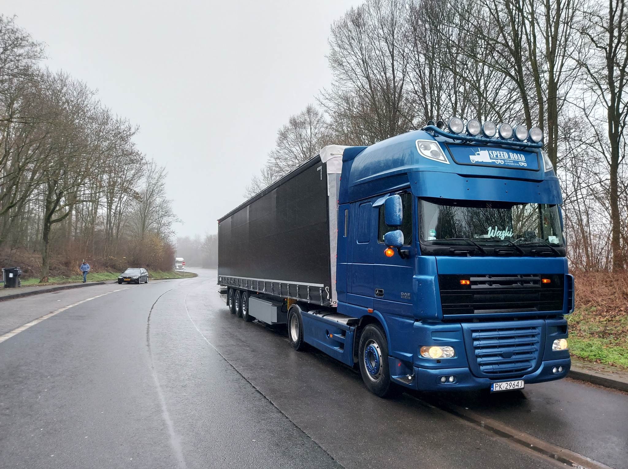 Błękitny ciągnik siodłowy DAF z czarną naczepą, tablica rejestracyjna PK 2964J, na mokrej drodze w jesiennej scenerii. Na dachu lampy.