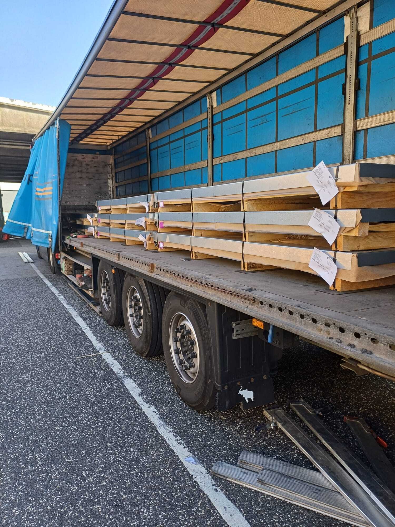 Ciężarówka z niebieską plandeką załadowana paletami z metalowymi elementami, zabezpieczonymi etykietami, widoczna z boku na parkingu.