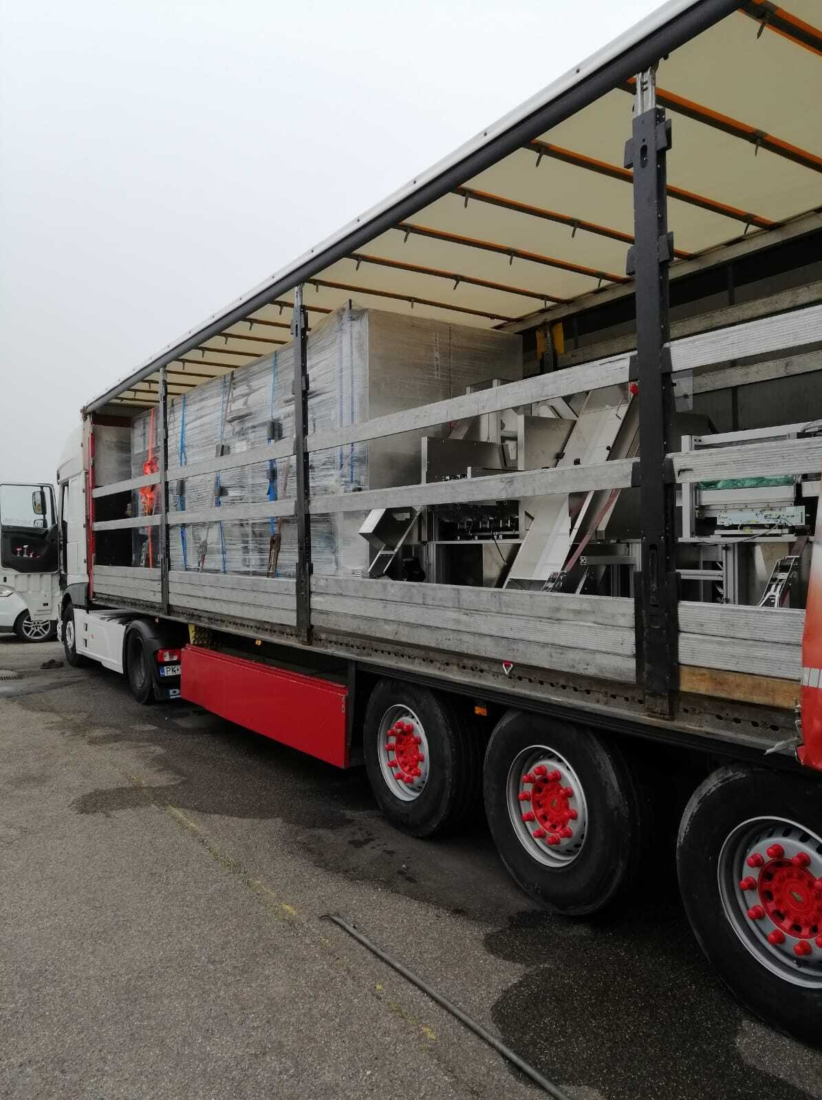 Ciężarówka z otwartą naczepą, przewożąca zabezpieczone folią maszyny przemysłowe. Widoczne czerwone akcenty na kołach i podwoziu. Transport ładunku w pochmurny dzień.