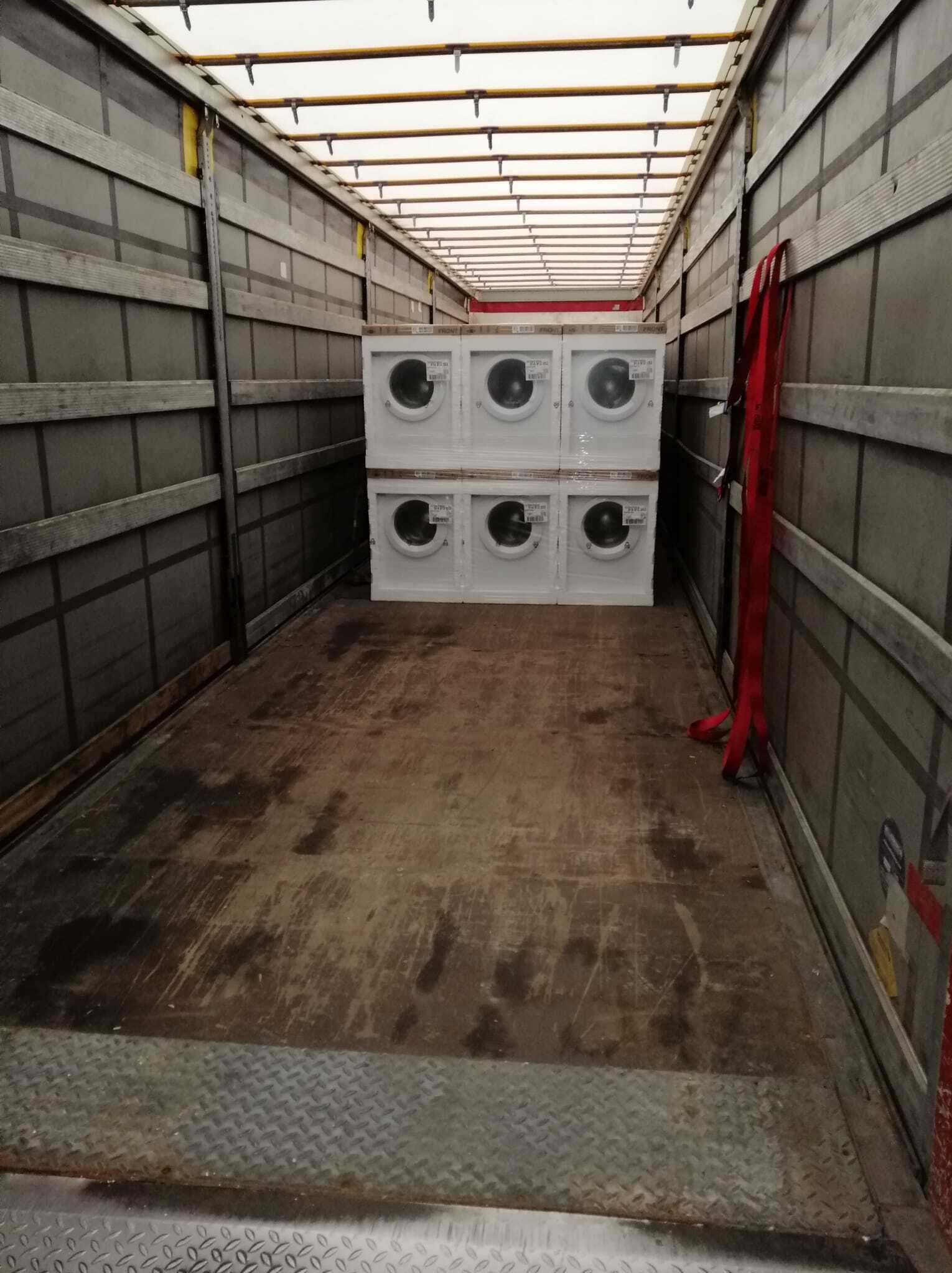 Wnętrze naczepy z sześcioma pralkami, zabezpieczonymi do transportu. Widoczne pasy mocujące i metalowa podłoga. Oświetlenie górne. Perspektywa z głębi naczepy.