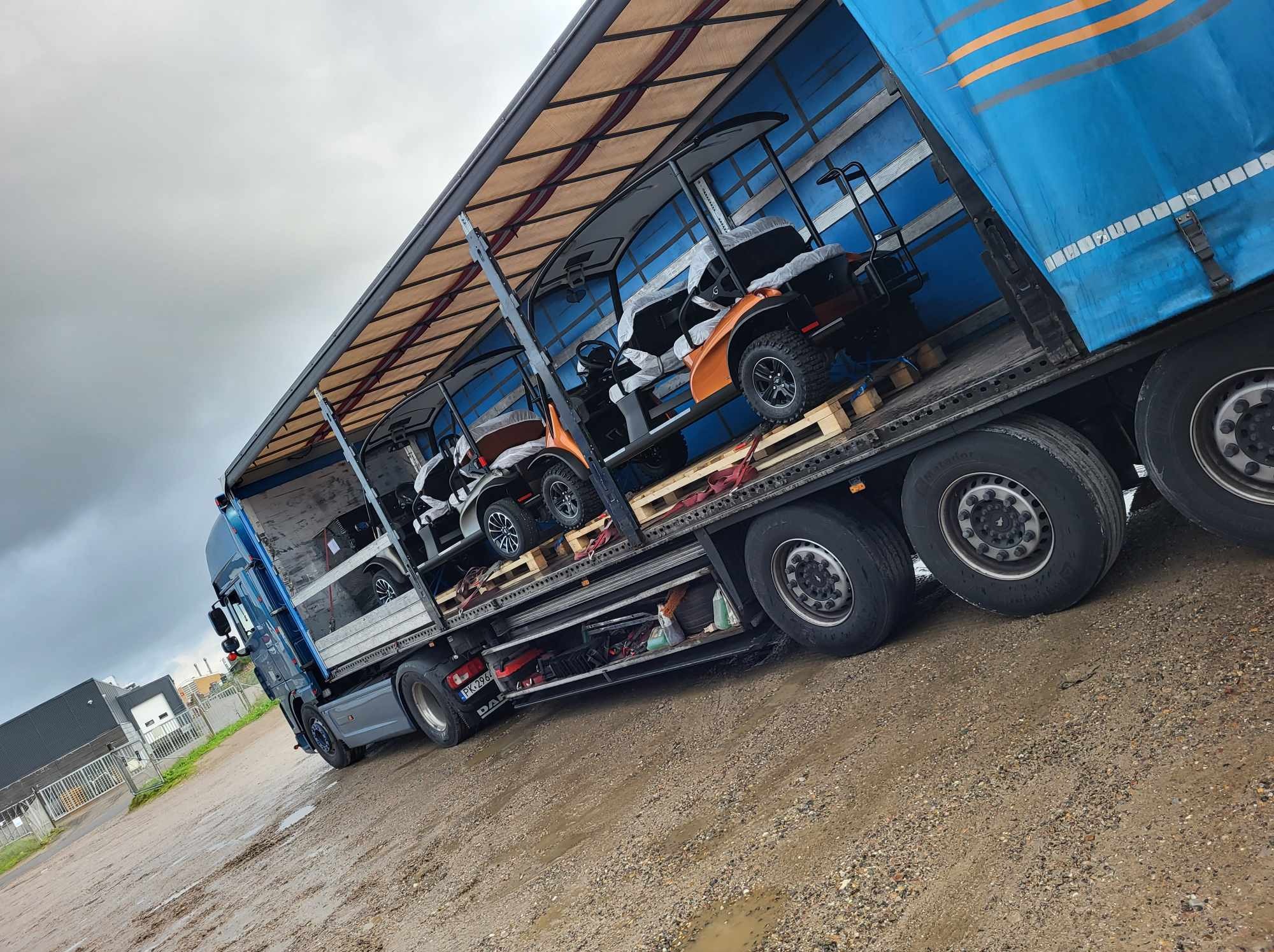 Ciężarówka z otwartą naczepą przewozi na paletach pomarańczowo-białe wózki golfowe. Widoczna boczna ściana naczepy i mokre podłoże.