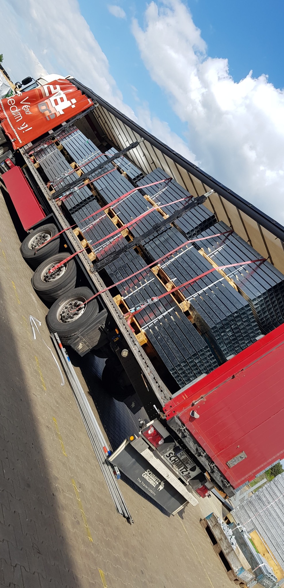 Otwarta naczepa ciężarówki załadowana paletami z metalowymi profilami, zabezpieczonymi czerwonymi pasami. Widoczne logo firmy transportowej i błękitne niebo.