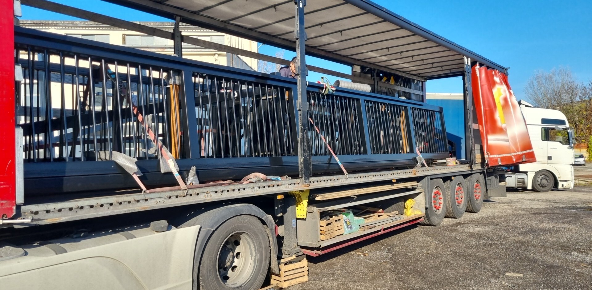 Ciężarówka z naczepą przewozi czarne, metalowe elementy ogrodzenia zabezpieczone pasami transportowymi w słoneczny dzień. Widoczne skrzynki i narzędzia.