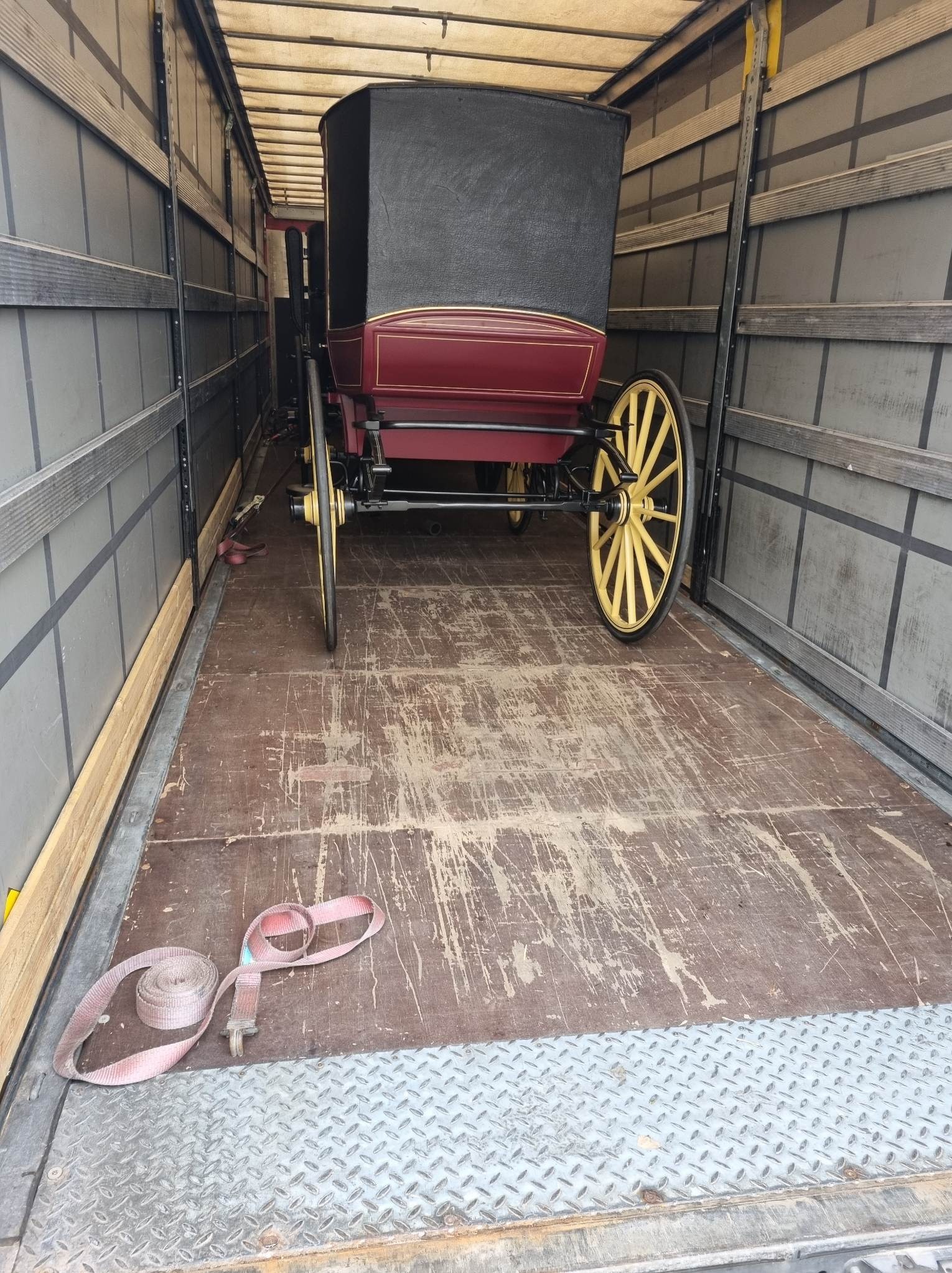 Wnętrze ciężarówki z zabytkowym powozem w kolorze bordowym i czarnym, zabezpieczonym pasami transportowymi. Podłoga drewniana, z widocznymi śladami użytkowania.