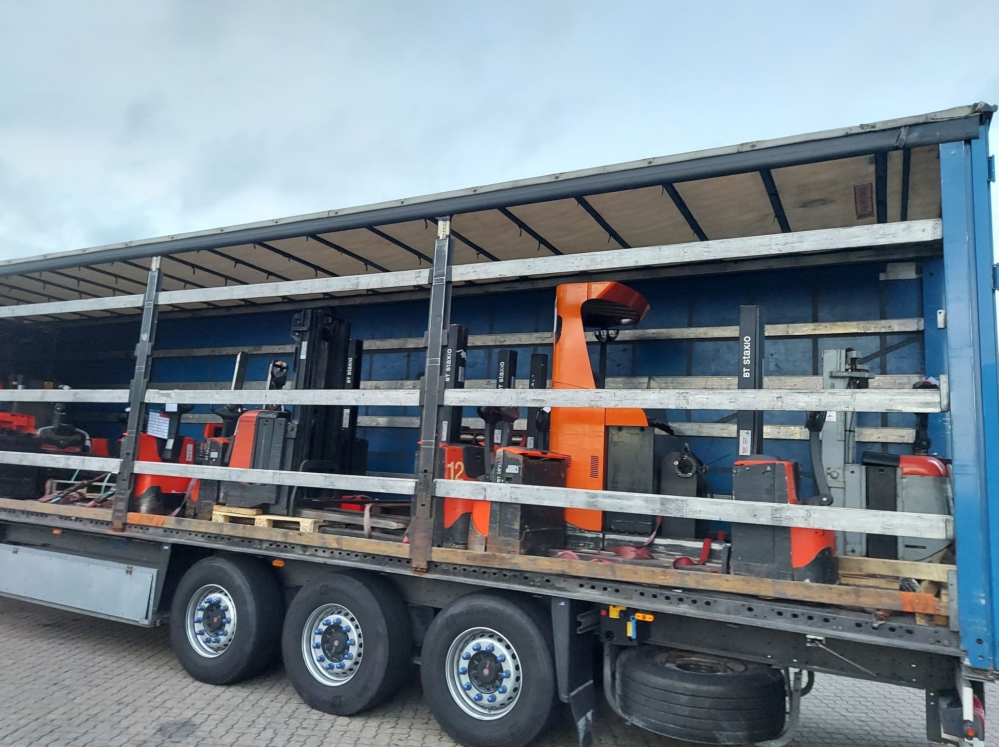 Ciężarówka z otwartą plandeką, wewnątrz kilka wózków widłowych zabezpieczonych drewnianymi belkami podczas transportu. Widoczne koła ciężarówki i fragment nawierzchni.