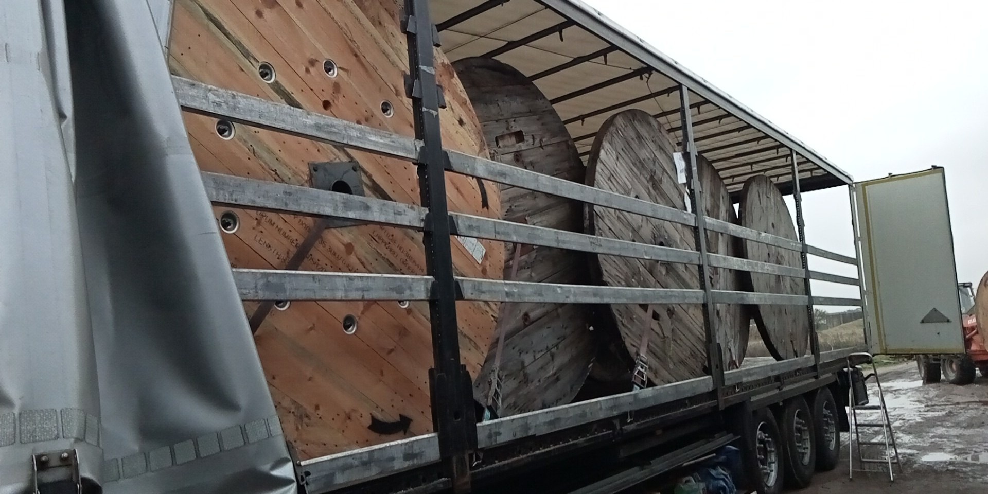 Otwarta naczepa z drewnianymi szpulami kablowymi, zabezpieczonymi metalowymi poprzeczkami. Widoczne detale konstrukcyjne naczepy i fragment terenu w tle.