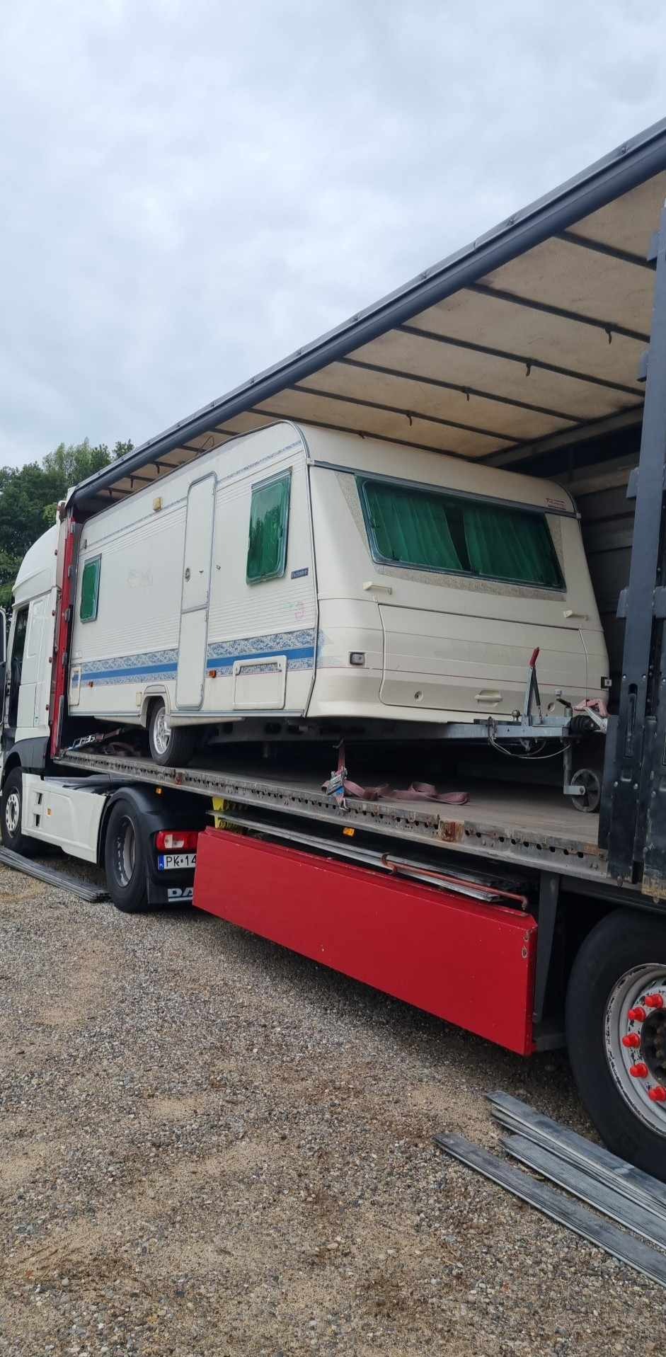 Transport przyczepy kempingowej na lawecie ciężarówki z czerwoną burtą. Widoczne zielone zasłony w oknach przyczepy i tablica rejestracyjna pojazdu. Szary, pochmurny dzień.