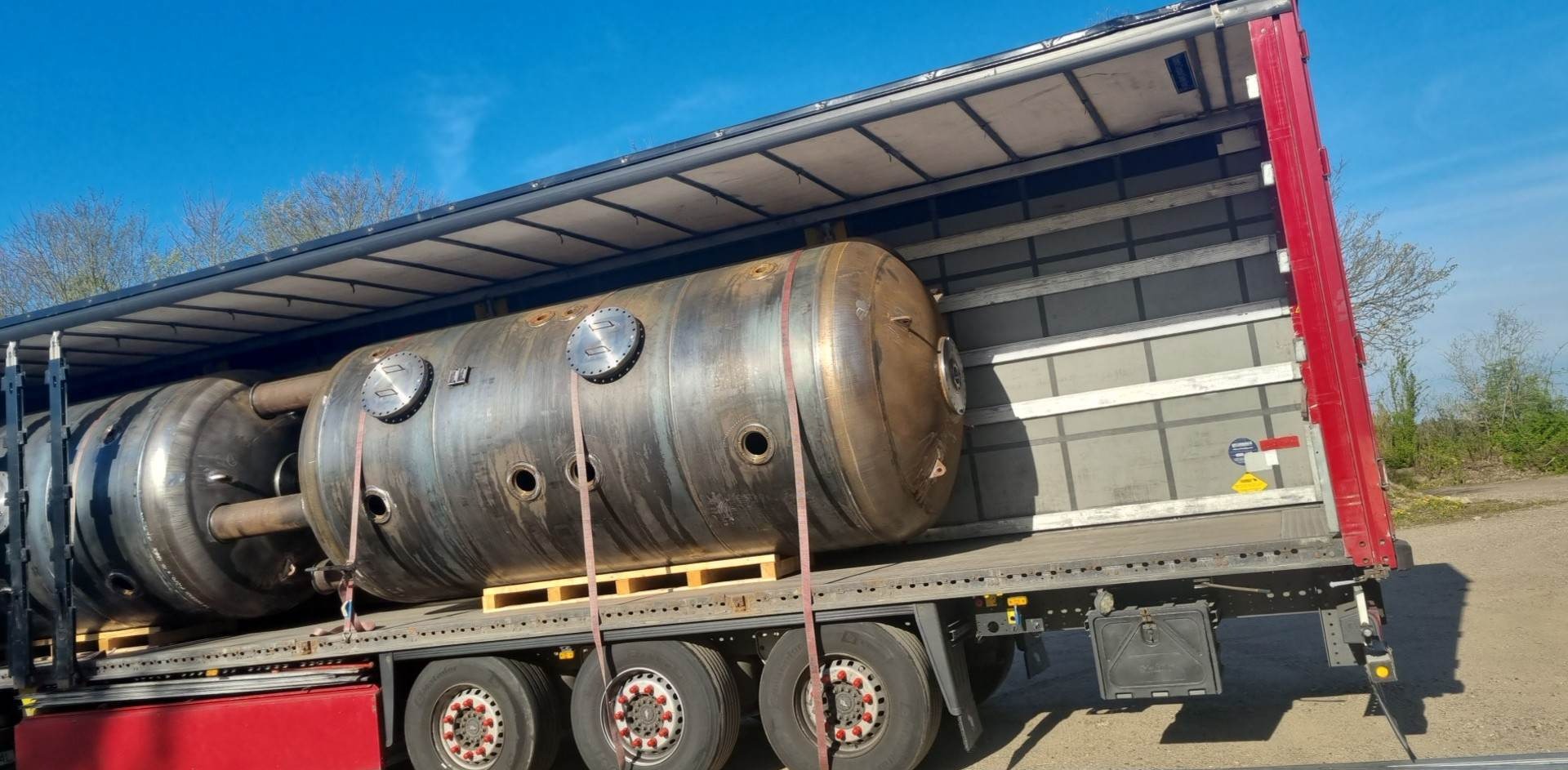 Ciężarówka z naczepą przewozi duże, metalowe zbiorniki. Zbiorniki zabezpieczone pasami, umieszczone na paletach. Błękitne niebo w tle.
