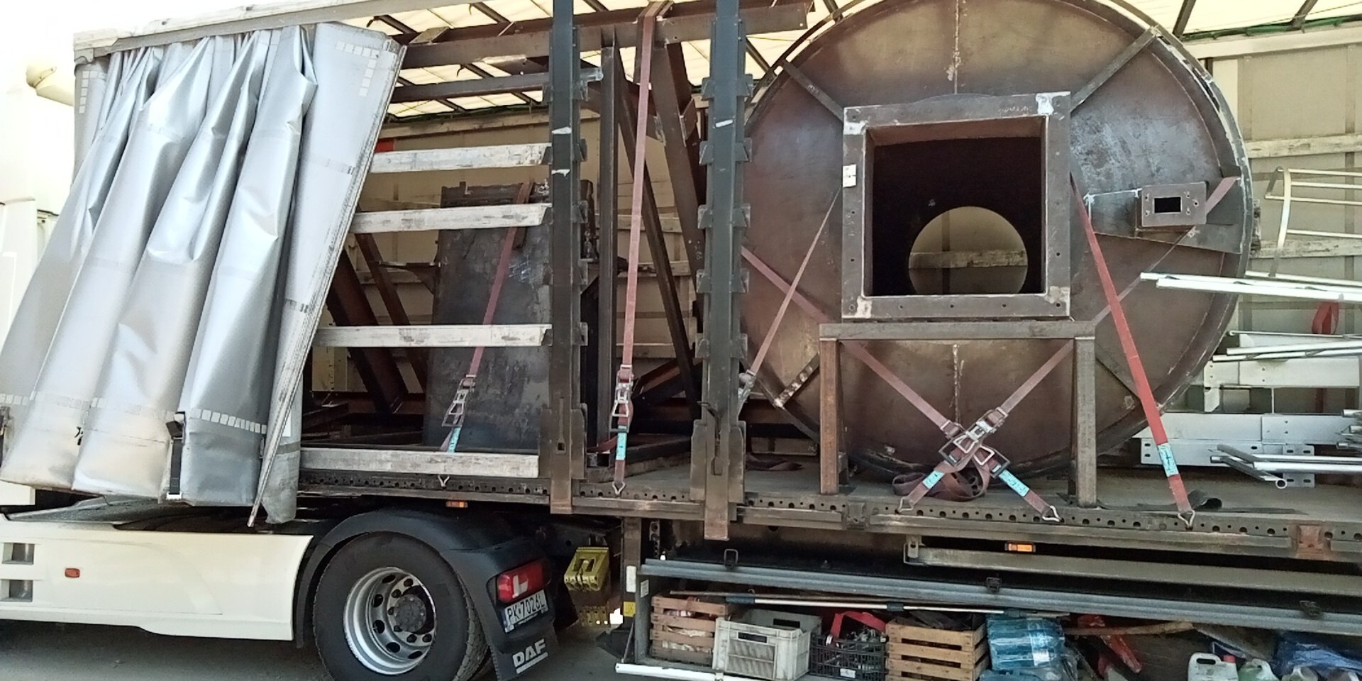 Transport specjalistyczny z Brzezin: ciężarówka z naczepą przewozi duży, okrągły element konstrukcyjny zabezpieczony pasami transportowymi. Widoczne skrzynki i narzędzia.