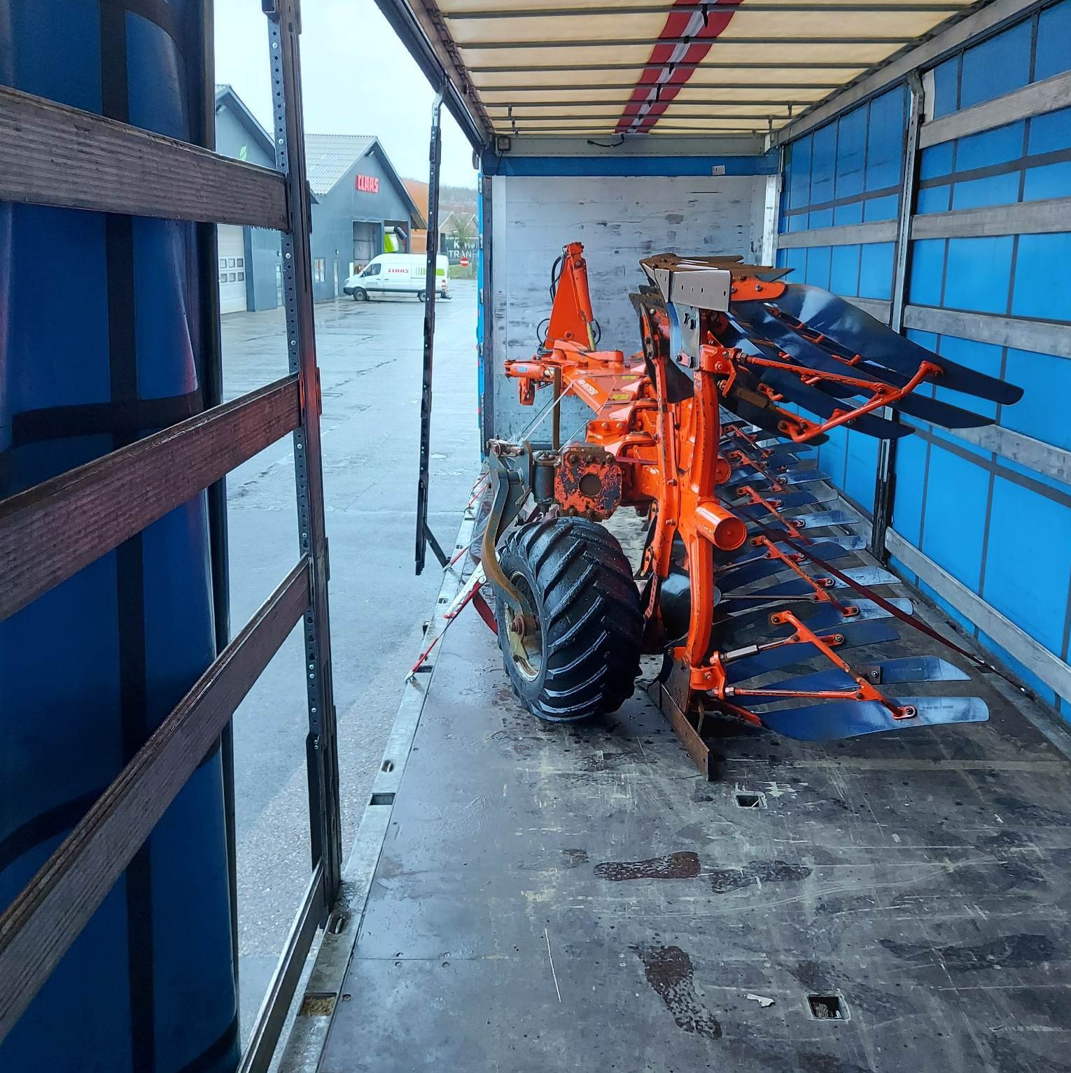 Wnętrze naczepy z pomarańczowym pługiem rolniczym. Widoczne mocowania i zabezpieczenia ładunku. W tle budynek firmy Claas i zaparkowany bus.