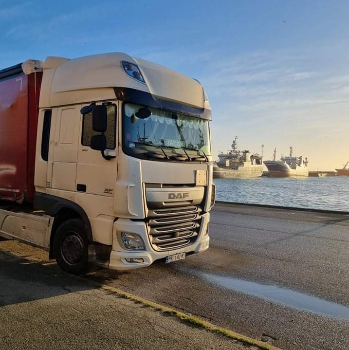 Ciężarówka DAF XF z naczepą na tle portu i statków, w słoneczny dzień. Widoczne refleksy na mokrej nawierzchni.
