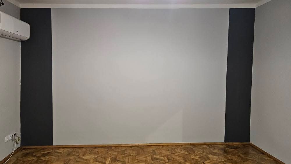 Ściana po malowaniu z ciemnymi pasami po bokach, parkiet i klimatyzator. Widoczne gniazdka elektryczne. Minimalistyczny wystrój wnętrza po renowacji.