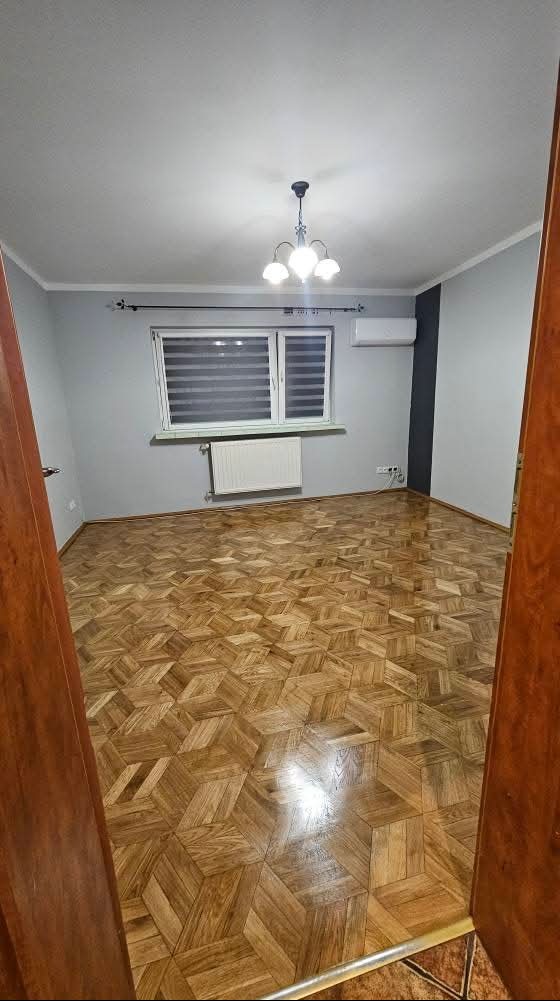 Pokój po remoncie: parkiet w geometryczny wzór, szare ściany z czarnym akcentem, białe okno z żaluzjami, grzejnik i klimatyzator. Widok zza drewnianej ościeżnicy.