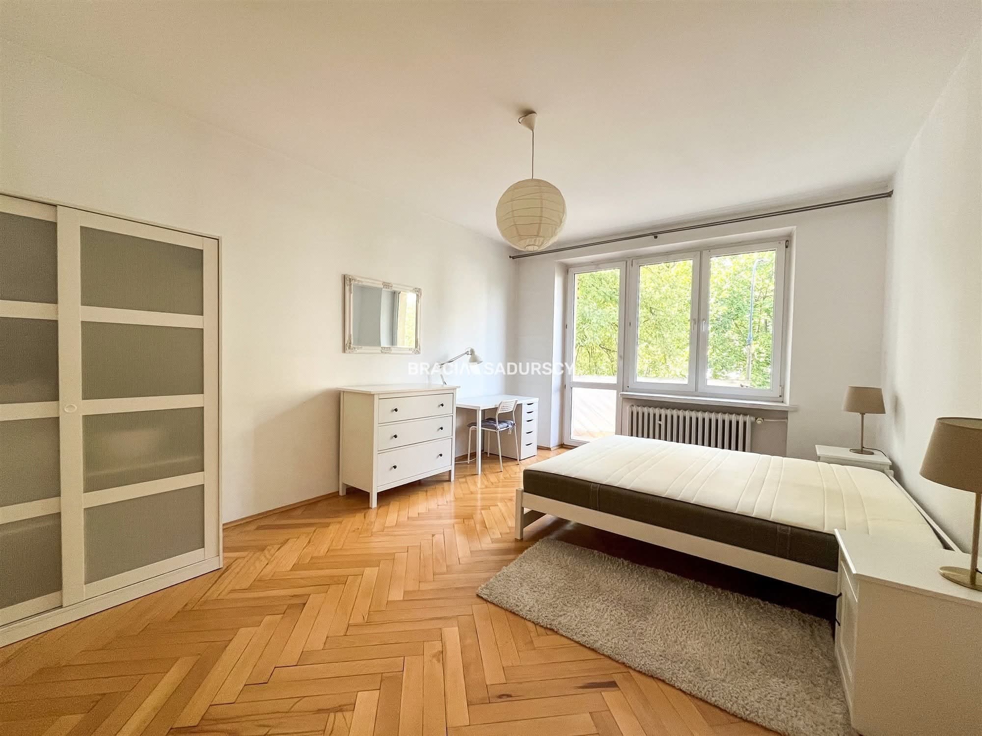 Jasne wnętrze sypialni z parkietem w jodełkę, białymi ścianami, meblami w stylu skandynawskim i dużym oknem z widokiem na zieleń. Minimalistyczny design.