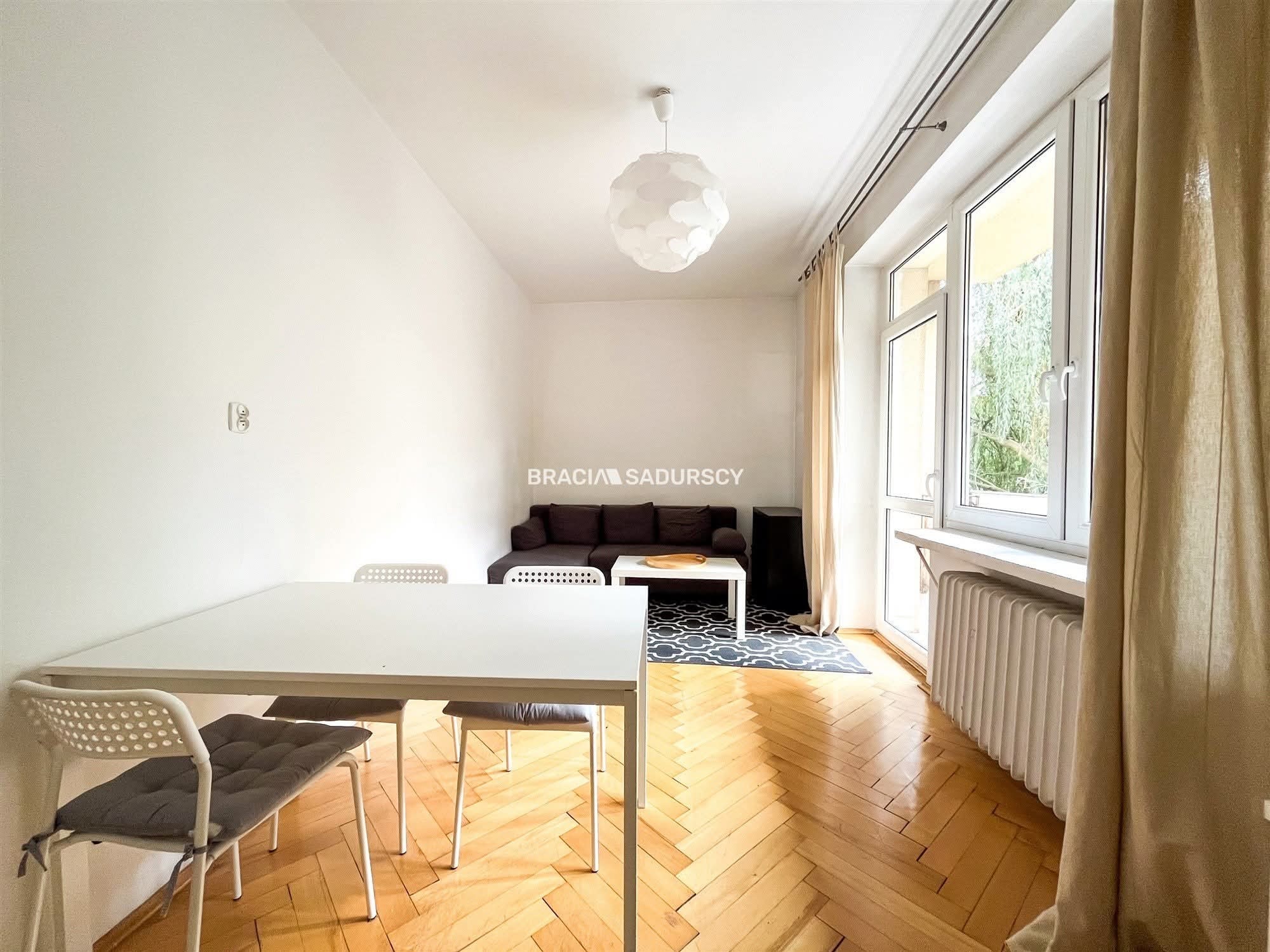 Jasne wnętrze po remoncie: parkiet w jodełkę, białe ściany, stół z krzesłami, sofa i okno z zasłonami. Widoczny grzejnik i lampa sufitowa. Minimalistyczny design.