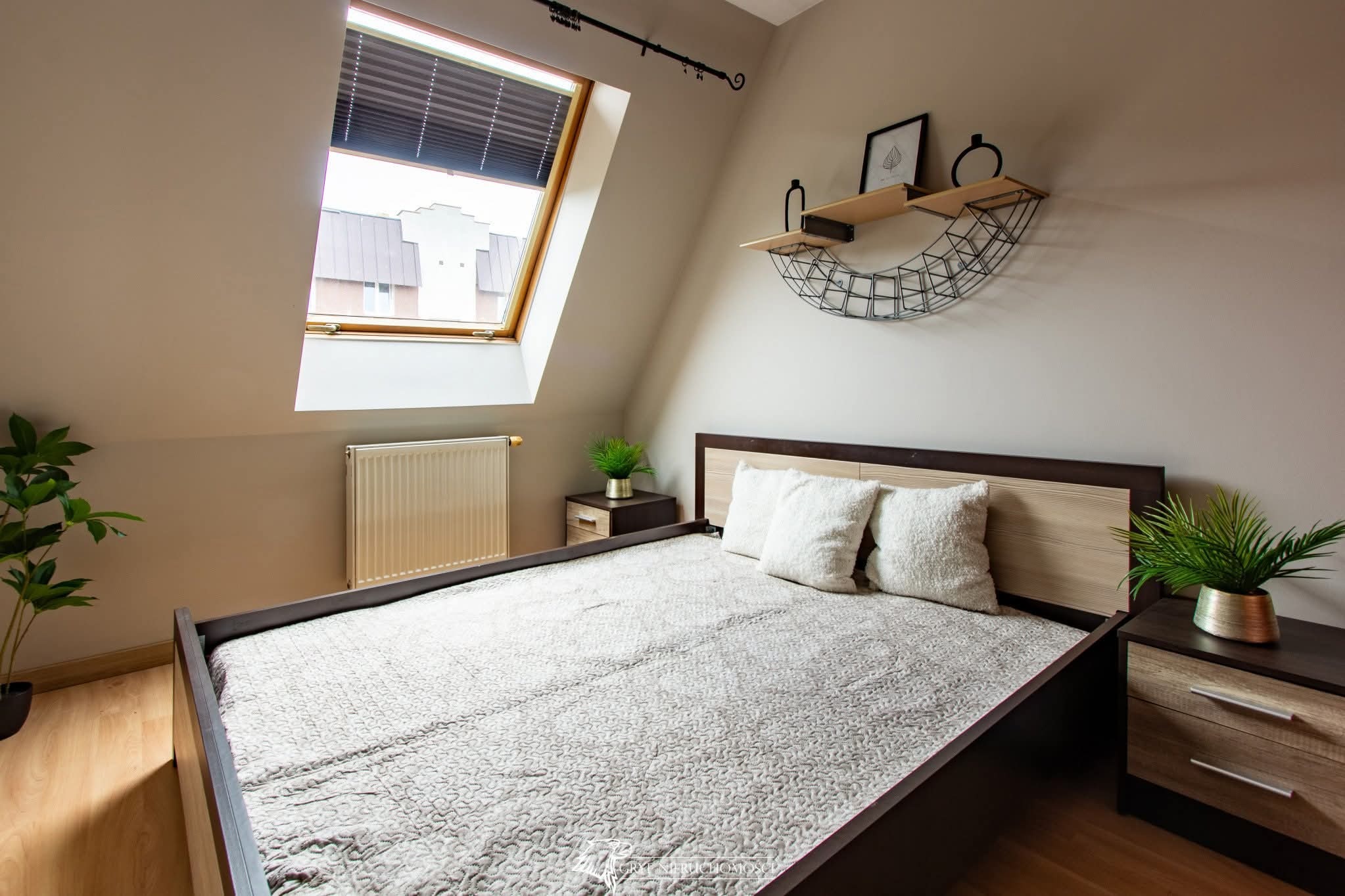 Beżowa sypialnia na poddaszu z oknem dachowym, dekoracyjną półką i roślinami doniczkowymi. Grzejnik pod oknem. Ustawiona pościel na łóżku.