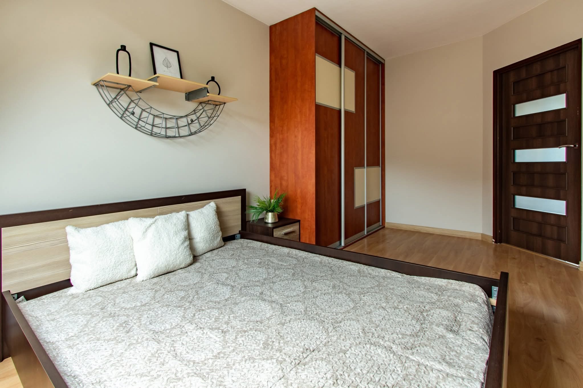 Jasne wnętrze sypialni z łóżkiem, drewnianą szafą przesuwną i półką dekoracyjną nad wezgłowiem. Parkiet na podłodze i ciemne drzwi po prawej stronie.