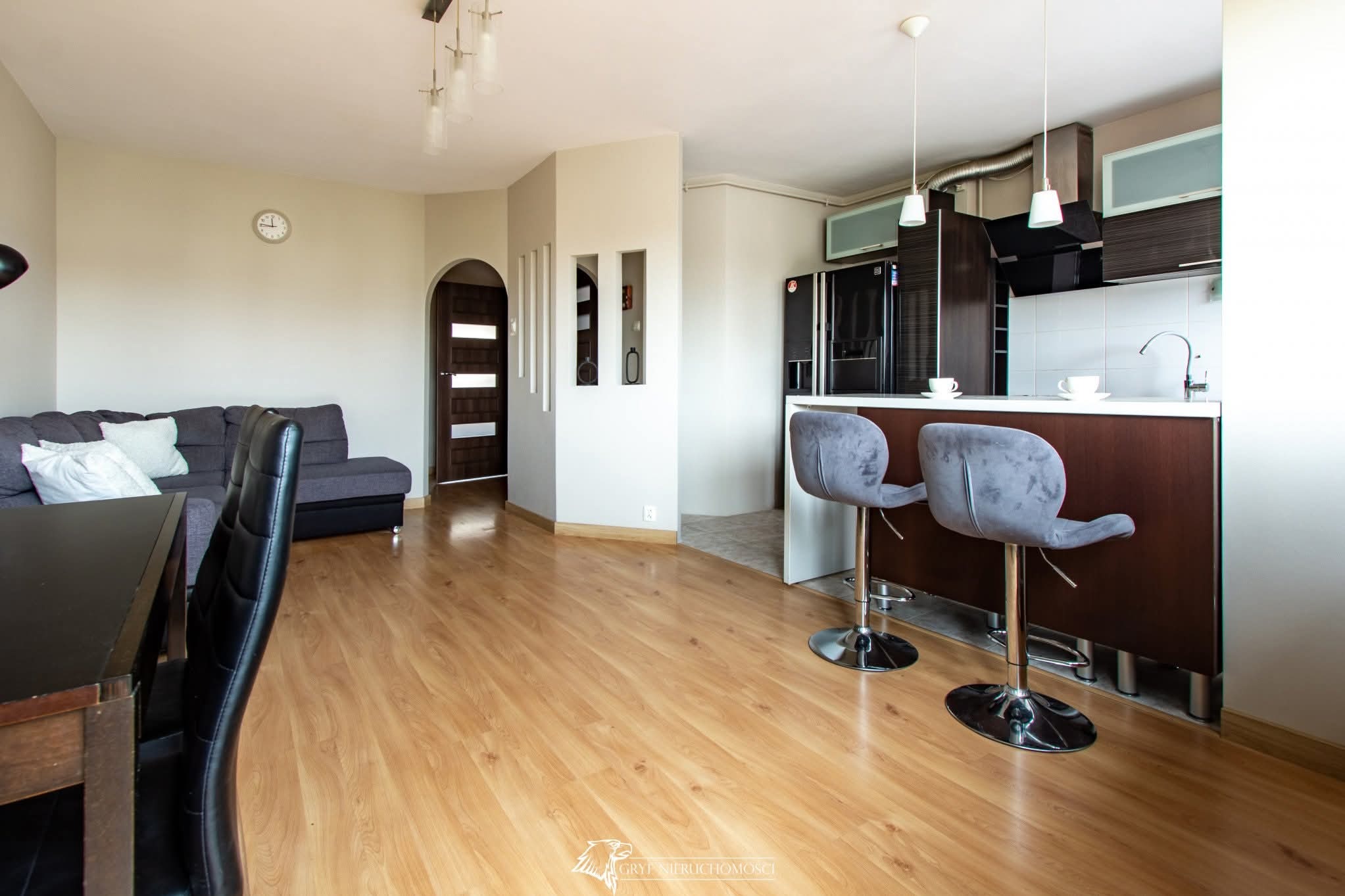 Jasne wnętrze mieszkania po remoncie: parkiet, pomalowane ściany, aneks kuchenny z barkiem i krzesłami. Widok na salon z szarą sofą i łukowate wejście do pokoju.