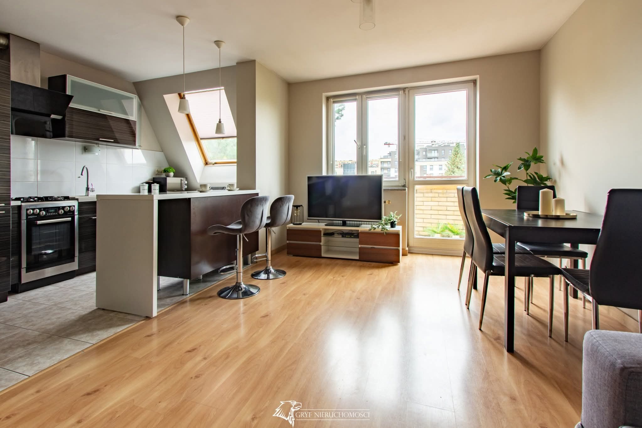 Nowoczesne wnętrze mieszkania z aneksem kuchennym, drewnianą podłogą i oknem dachowym. Widoczny stół z krzesłami i telewizor. Jasne, przestronne pomieszczenie.