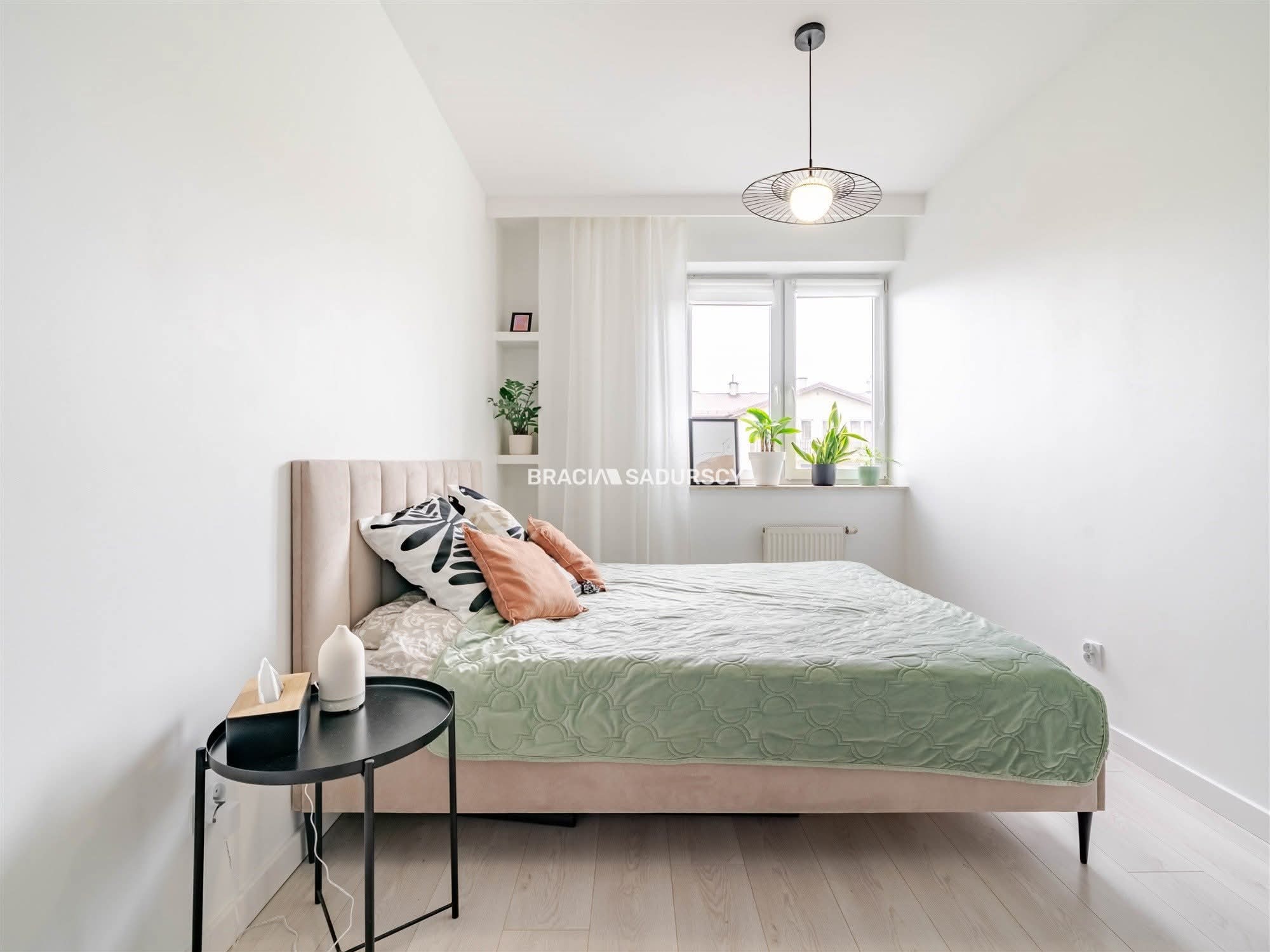 Jasna sypialnia z beżowym łóżkiem, zieloną narzutą i okrągłym stolikiem. Białe ściany i podłoga, lampa wisząca nad łóżkiem. Minimalistyczny wystrój.