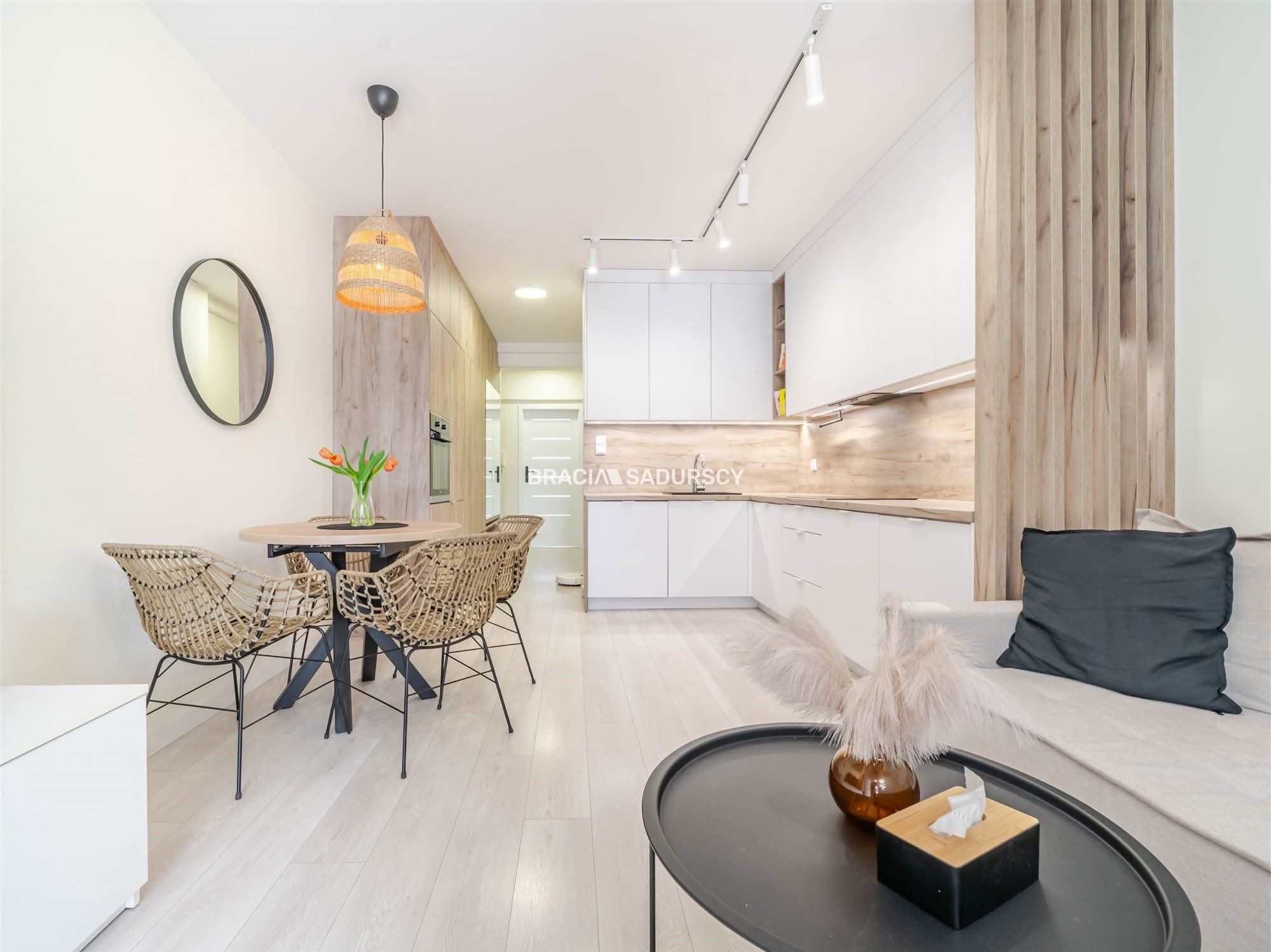 Jasne wnętrze mieszkania po remoncie: biała kuchnia z drewnianym blatem, okrągły stół z plecionymi krzesłami, drewniane lamele na ścianie i oświetlenie punktowe.