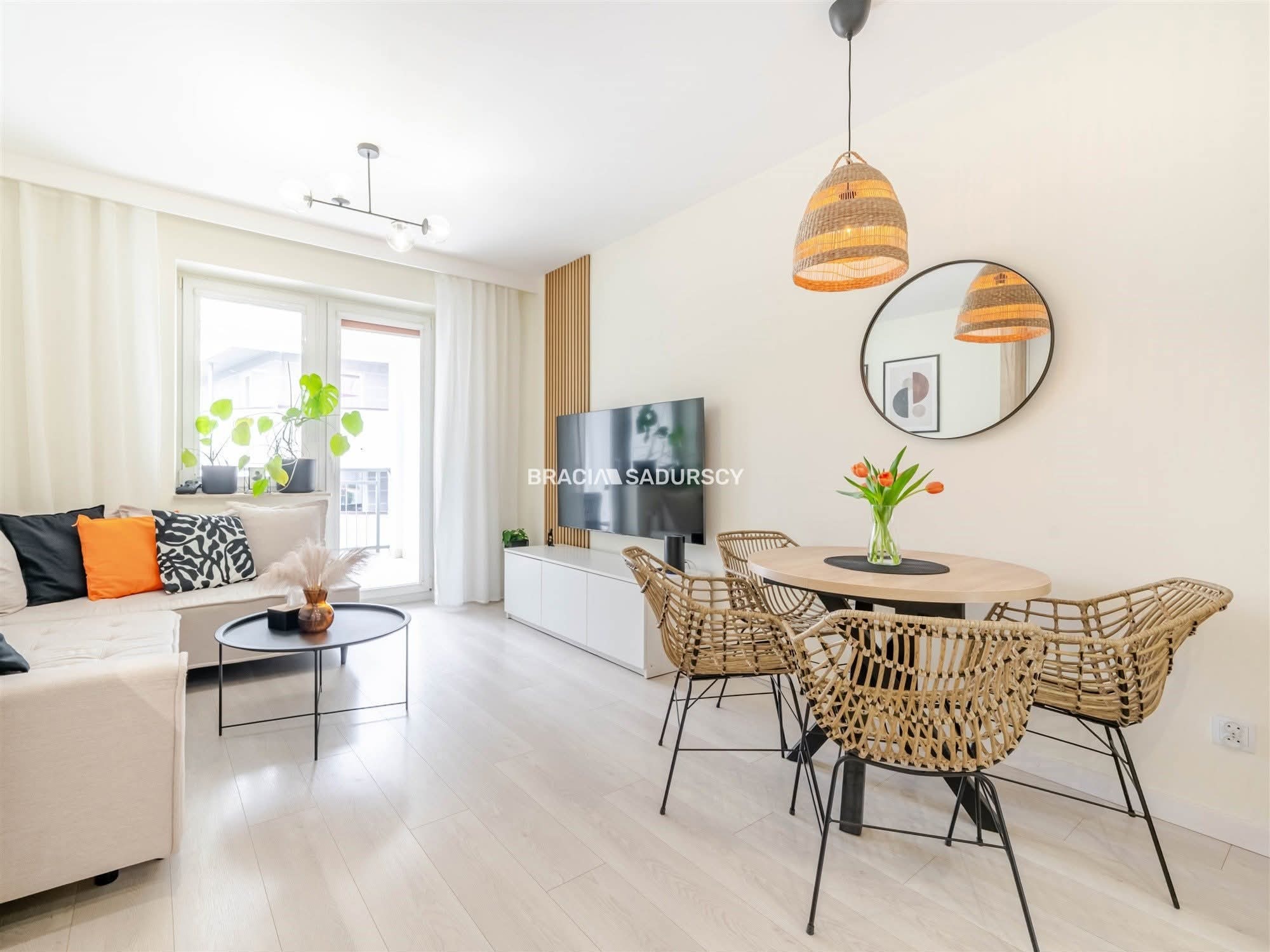 Jasne wnętrze: salon z sofą, stolikiem, TV i okrągłym stołem z krzesłami z wikliny. Na ścianie okrągłe lustro i lampa z abażurem. Parkiet w odcieniach bieli.