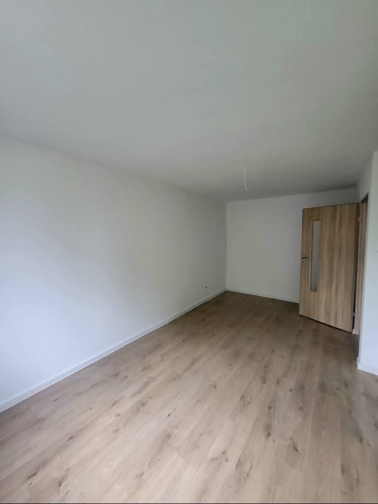 Jasne, puste wnętrze po remoncie: białe ściany, parkiet w odcieniach beżu i drewniane drzwi z przeszkleniem. Wystający kabel z sufitu.