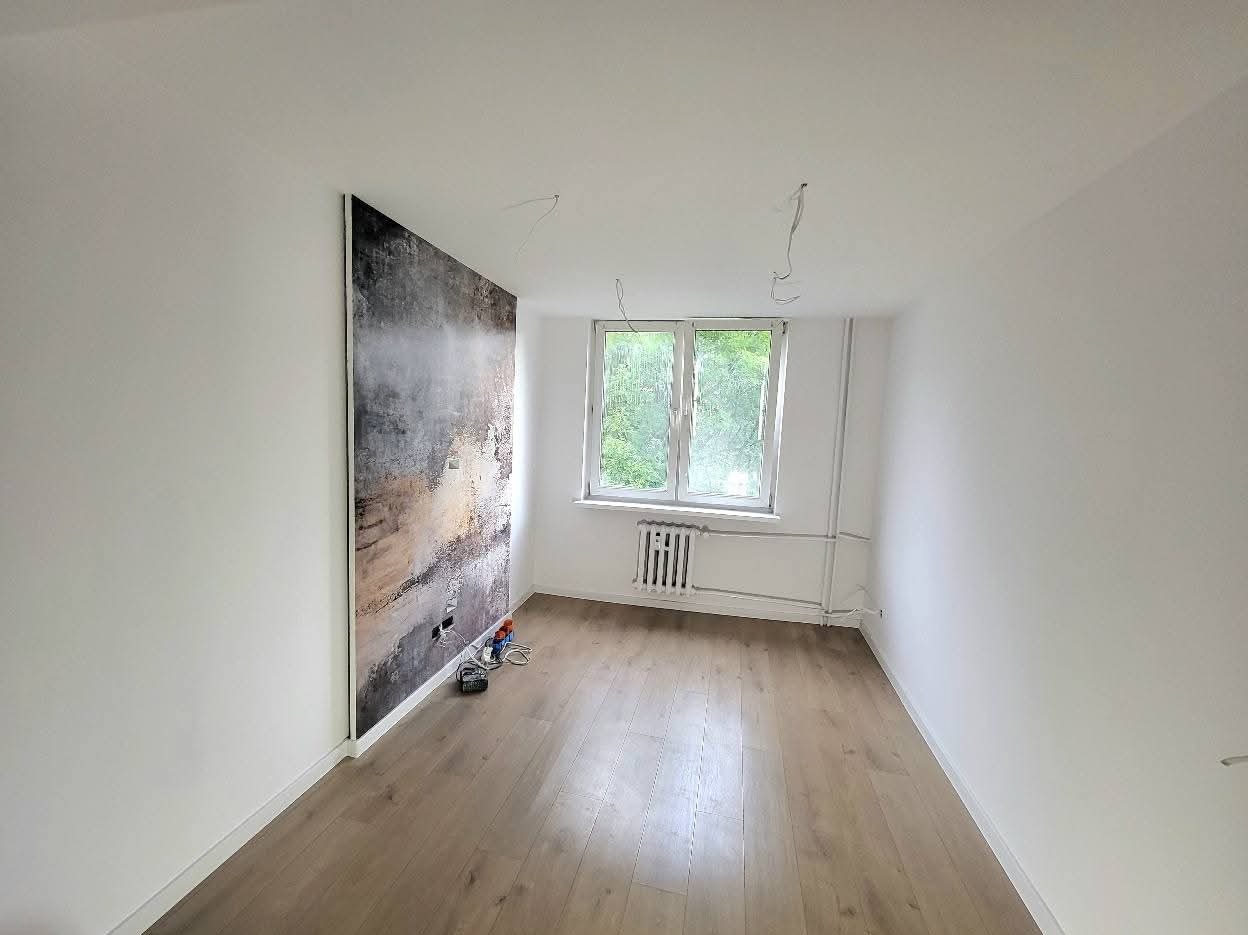 Pokój po remoncie: jasne ściany, drewniana podłoga, okno z widokiem na zieleń, na jednej ze ścian dekoracyjny panel imitujący abstrakcyjny obraz.