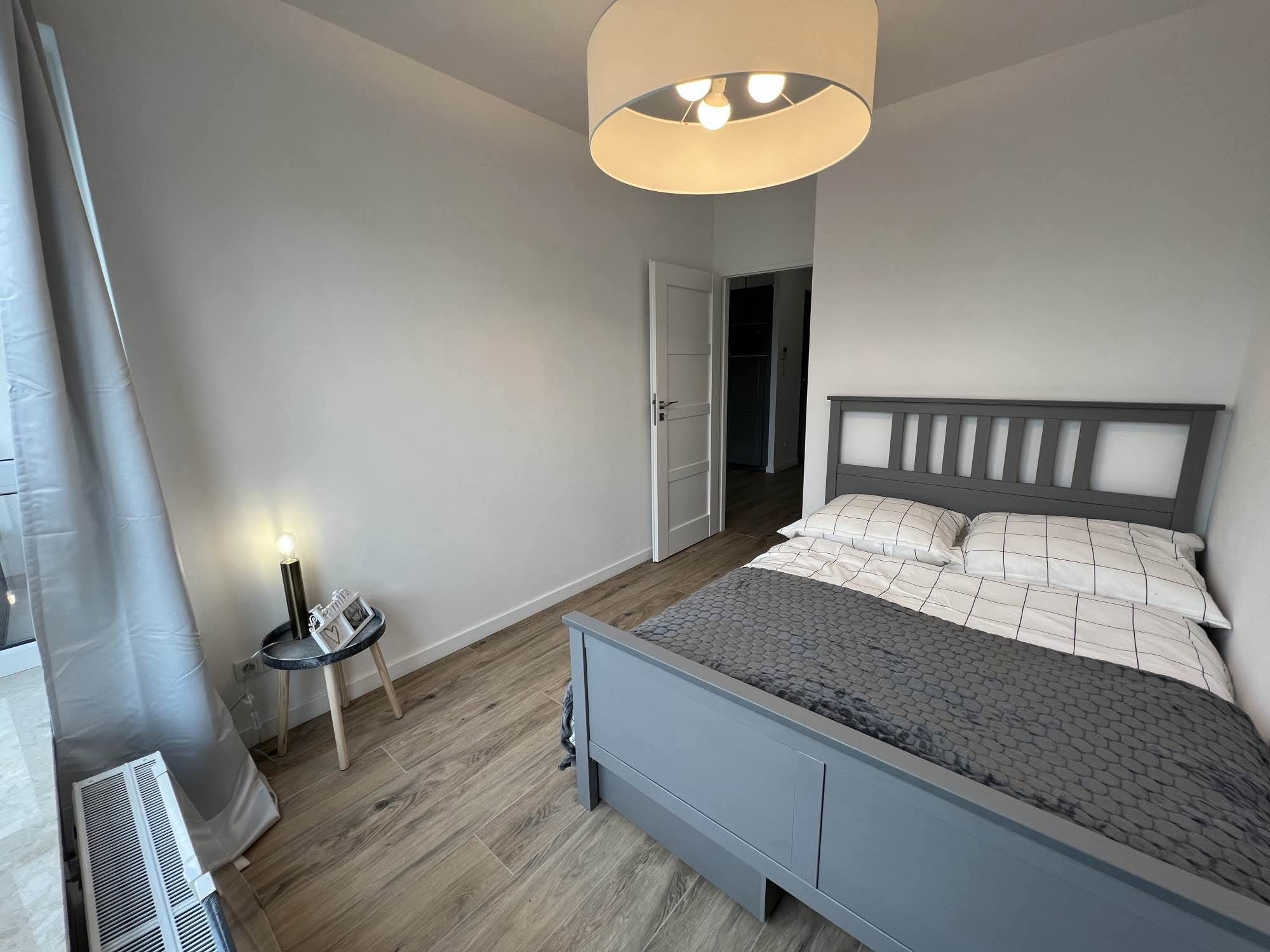 Jasne wnętrze sypialni: szare łóżko z wzorzystą pościelą, drewnopodobne panele, białe ściany i okrągła lampa sufitowa. Subtelne oświetlenie i minimalistyczny wystrój.