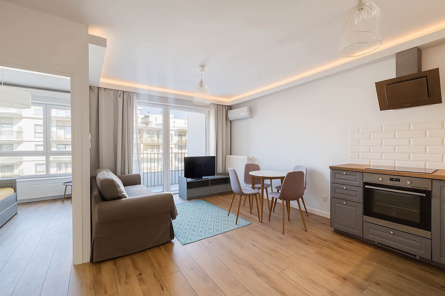 Jasne wnętrze mieszkania po wykończeniu: drewniana podłoga, szare meble kuchenne i wypoczynkowe, białe ściany z oświetleniem LED. Widok na balkon.