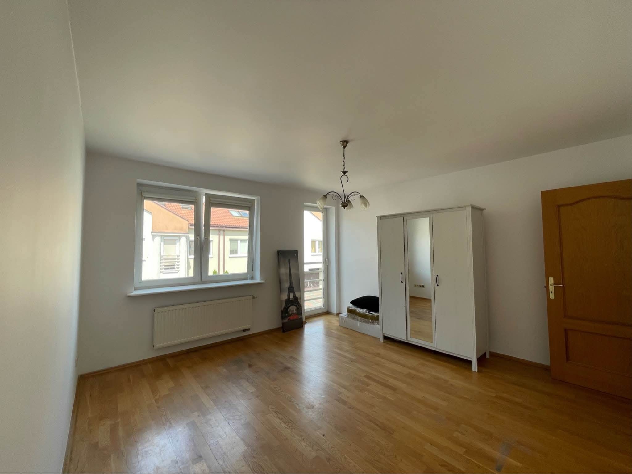 Jasne wnętrze pokoju z parkietem i białymi ścianami po malowaniu. Widoczne okno, grzejnik, szafa z lustrami i plakat z wieżą Eiffla.