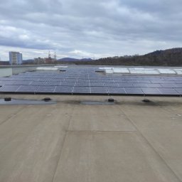 GLOBAL SOLUTIONS SERHII RAKOMIN - Instalacja fotowoltaiczna na dachu w Sosnowcu. Widoczne panele słoneczne, świetliki dachowe i sylwetka osoby na tle zabudowy miejskiej i zalesionych wzgórz.