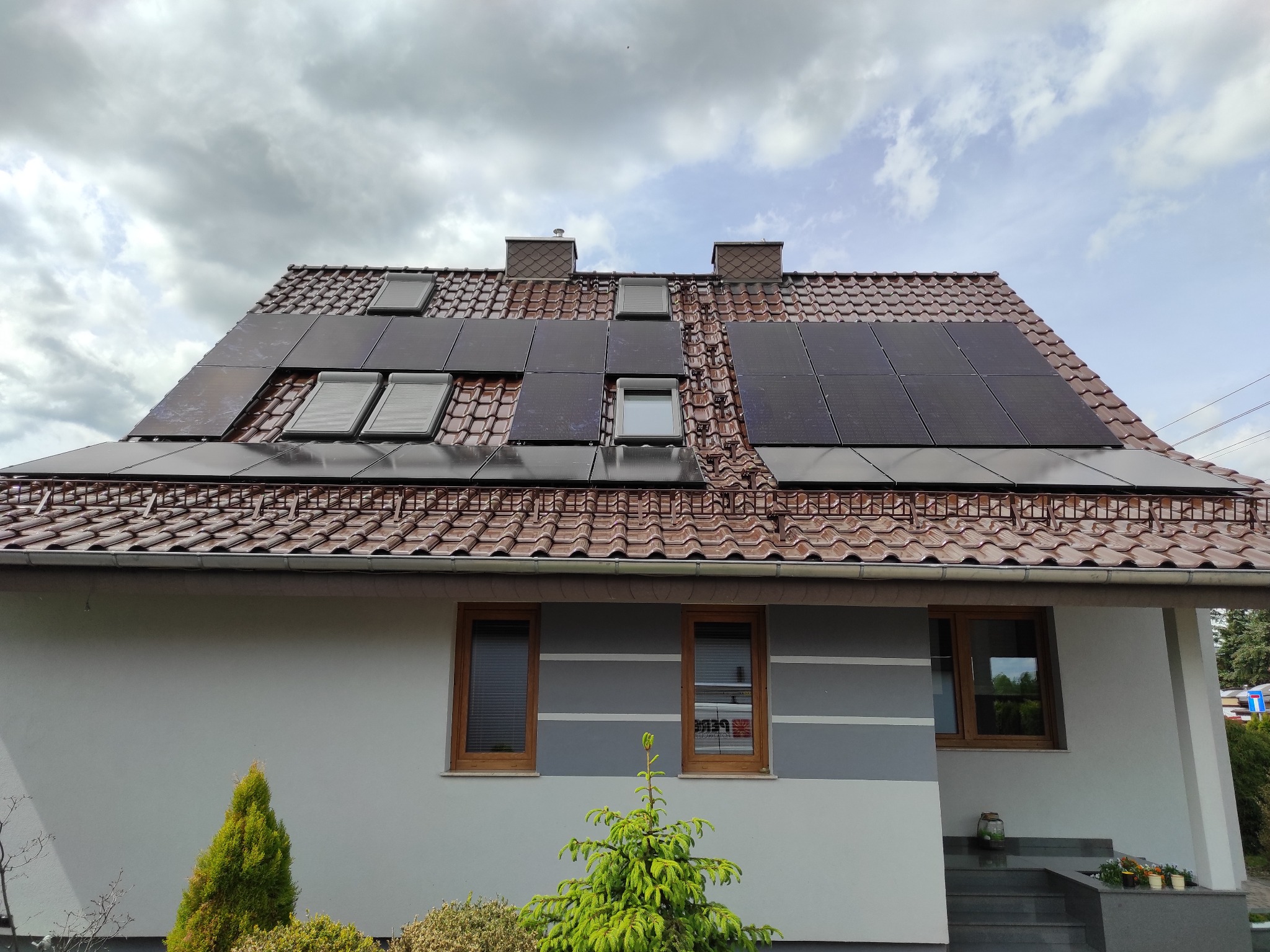 Dom z panelami fotowoltaicznymi na dachu z brązowej dachówki, zamontowanymi między oknami dachowymi. Szara fasada z brązowymi oknami i dekoracyjnymi pasami.