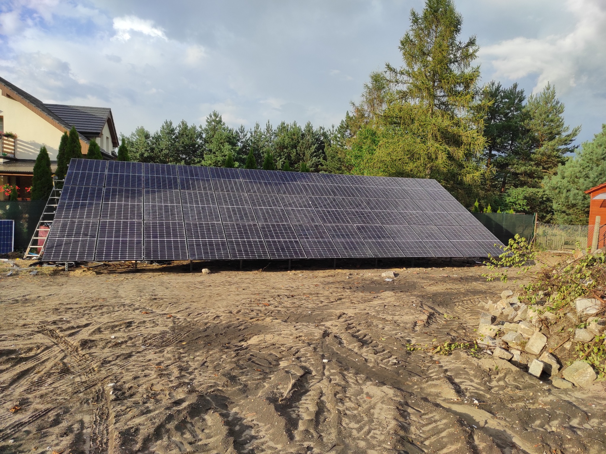 Rozległa instalacja fotowoltaiczna na gruncie, z drabiną opartą o panele. W tle dom z czerwonymi kwiatami na balkonie i wysokie drzewa pod pochmurnym niebem.