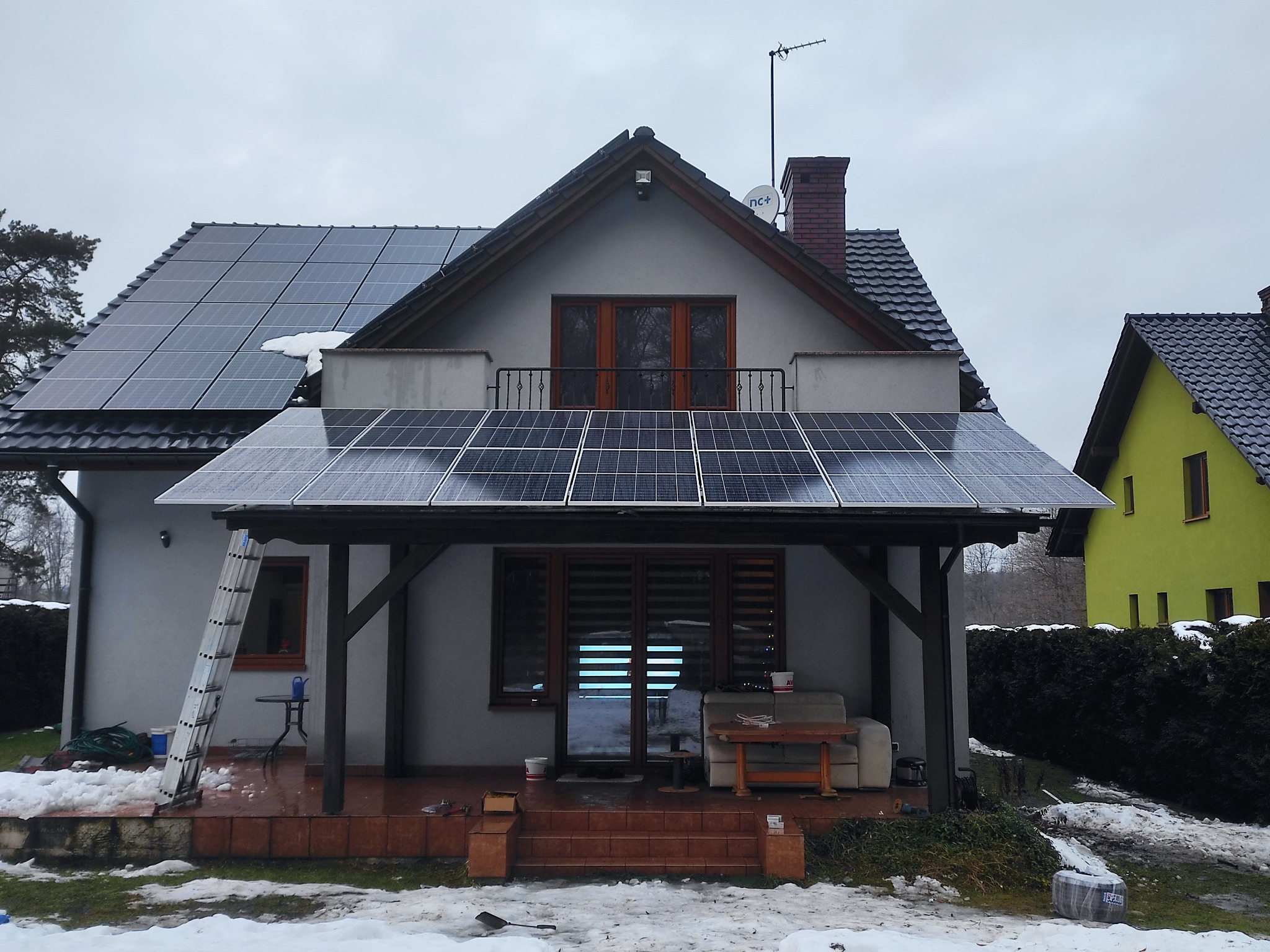 Dom z panelami fotowoltaicznymi na dachu i zadaszeniu tarasu, drabina oparta o ścianę. Zimowy krajobraz z resztkami śniegu. Pochmurne niebo.