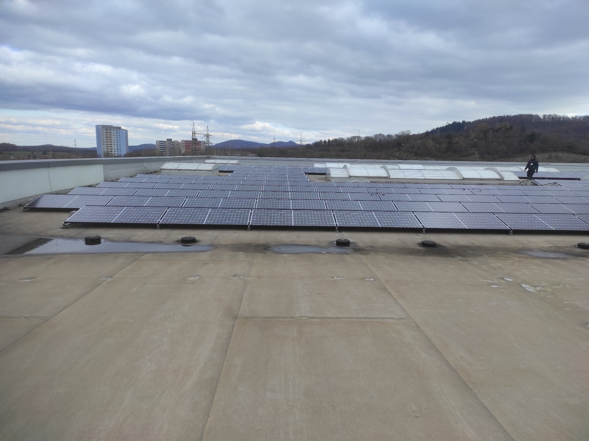 Instalacja fotowoltaiczna na dachu w Sosnowcu. Widoczne panele słoneczne, świetliki dachowe i sylwetka osoby na tle zabudowy miejskiej i zalesionych wzgórz.