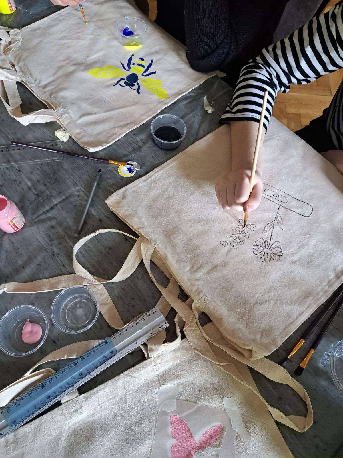 Dzieci malują farbami na bawełnianych torbach, tworząc unikalne wzory motyli, kwiatów i owadów. Widoczne pędzle, farby i linijki.