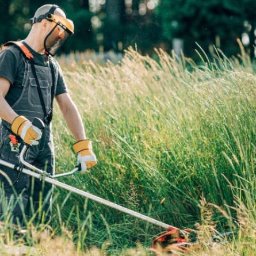 Marek Lubczyński - Mężczyzna w kombinezonie, z osłoną twarzy i w rękawicach, kosi wysoką trawę za pomocą podkaszarki spalinowej. Słoneczny dzień, rozproszone światło.