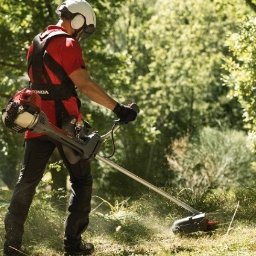 Marek Lubczyński - Pracownik w kasku i z kosą spalinową Honda, tnący trawę na tle zieleni. Ujęcie z boku, z widocznym pyłem i świeżo skoszoną trawą.