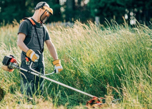 Mężczyzna w kombinezonie, z osłoną twarzy i w rękawicach, kosi wysoką trawę za pomocą podkaszarki spalinowej. Słoneczny dzień, rozproszone światło.