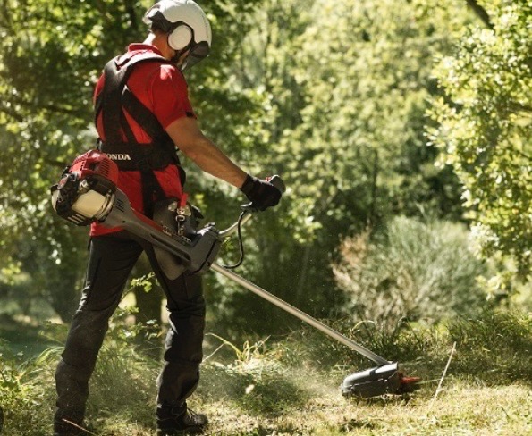 Pracownik w kasku i z kosą spalinową Honda, tnący trawę na tle zieleni. Ujęcie z boku, z widocznym pyłem i świeżo skoszoną trawą.
