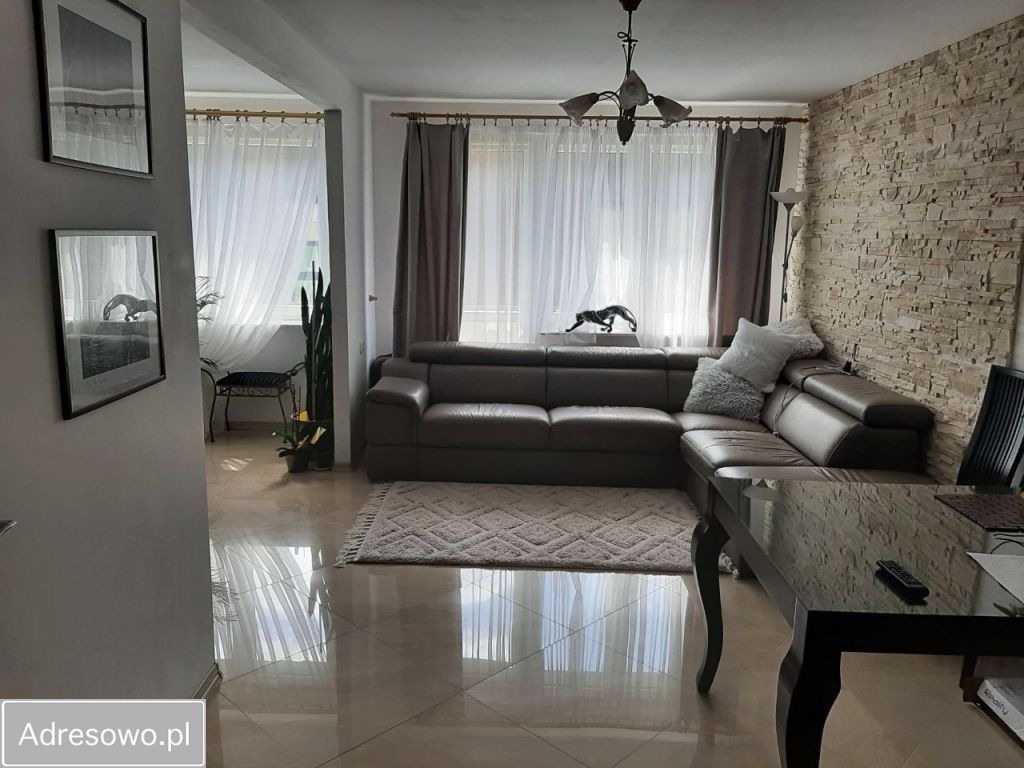 Jasne wnętrze salonu z narożną sofą, kamienną ścianą i błyszczącą podłogą. Widoczne dekoracje: lampa, zasłony, obrazy na ścianie. Uwagę zwraca czystość i porządek.