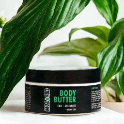 Patrycja Makles - Słoik z masłem do ciała CBD na białym tle, otoczony zielonymi liśćmi roślin. Etykieta z napisem 'Body Butter' i informacjami o składnikach. Stonowana kolorystyka, naturalne światło.