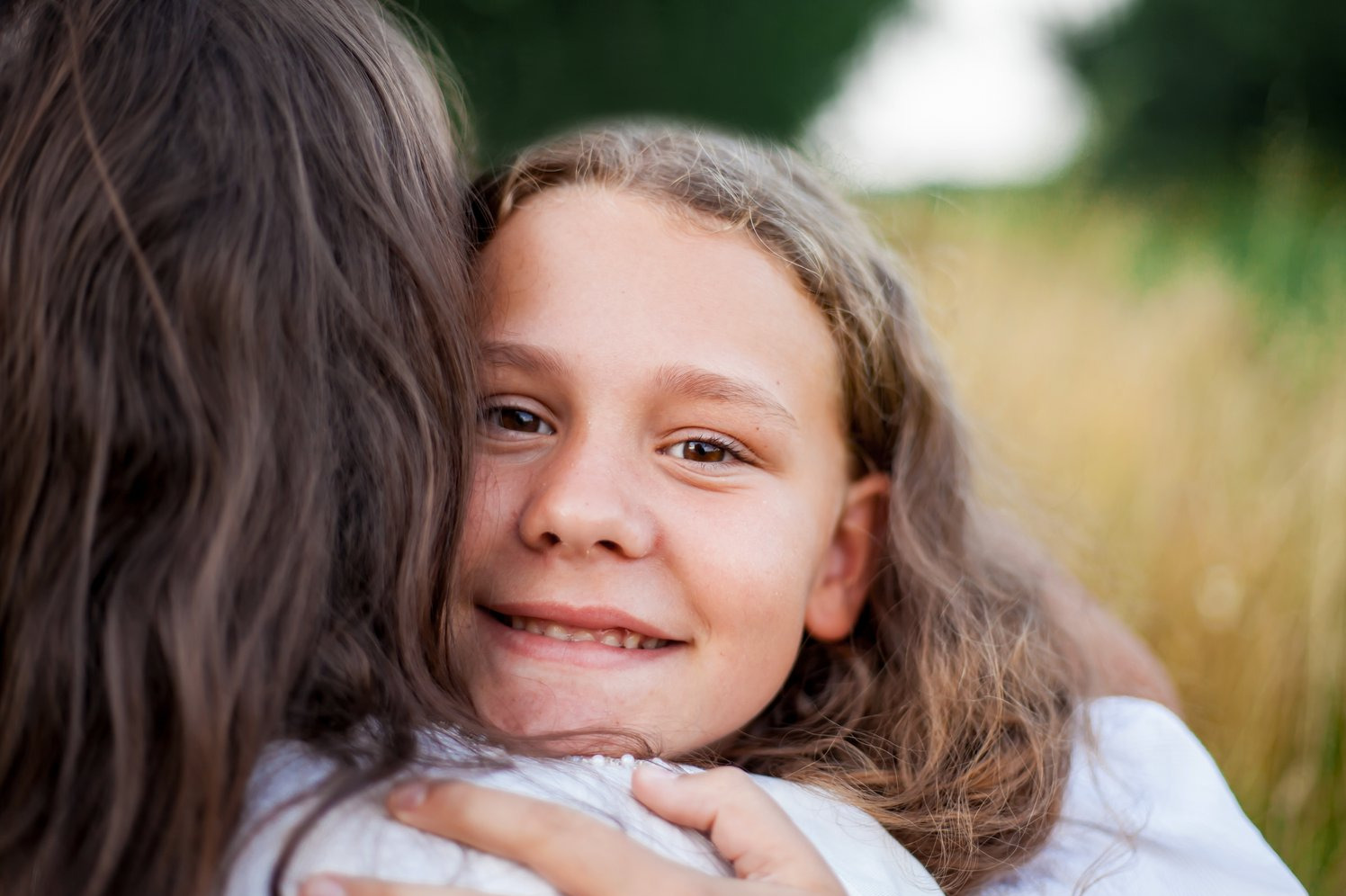 Uśmiechnięta dziewczynka z brązowymi włosami przytula drugą osobę na tle łąki, ciepłe światło, sesja rodzinna w plenerze.